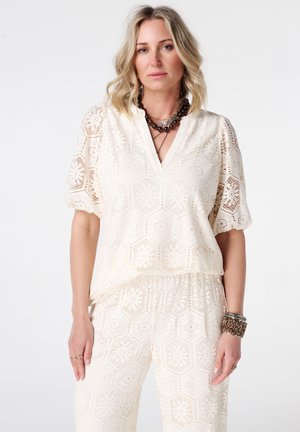 Femme aux cheveux blonds portant un chemisier blanc à motif en dentelle et un pantalon assorti, accessoirisée de colliers et bracelets superposés.