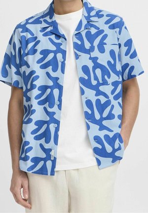 Homme portant une chemise à manches courtes bleu clair avec un motif abstrait de feuilles bleu foncé par-dessus un T-shirt blanc uni.