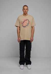 Bežs pārspīlēts T-krekls ar sarkanu un melnu tekstu un riņķveida ērkšķu dizainu; saskaņots ar melnām cargo biksēm un baltām kedām.