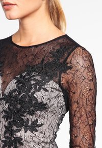 Svart spetsblus med blommönster broderat vid barmen och genomskinliga långärmar, som visar upp intrikata mönster och texturerade detaljer.