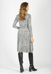 Szara sukienka z długim rękawem z abstrakcyjnym czarno-białym wzorem, zebrana w talii, zestawiona z czarnymi kozakami sięgającymi kolan, widziana od tyłu.