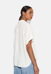 White short-sleeve blouse featuring a collar, soft texture, and a relaxed fit, with rounded hemline and back seam detail.