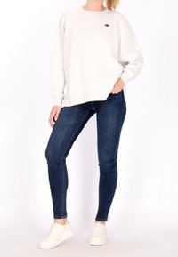 Vaaleanharmaa huppari, jossa on huomaamaton tummansininen logo, rento malli, yhdistettynä tummansinisiin kapeisiin farkkuihin ja valkoisiin lenkkareihin.