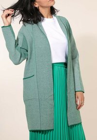 Cardigan long rayé vert et blanc avec un front ouvert et deux poches, porté sur une jupe plissée verte et un haut blanc. Texture lisse.