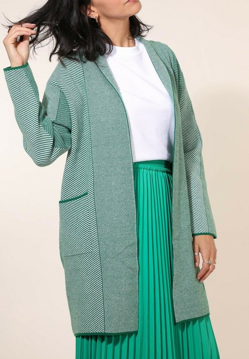 Cardigan long rayé vert et blanc avec un front ouvert et deux poches, porté sur une jupe plissée verte et un haut blanc. Texture lisse.