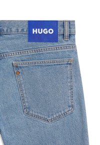 Sinised teksapüksid, millel on ristkülikukujuline sinine silt tekstiga "HUGO." Taga tasku ja kontrastsed oranžid õmblused. Pehme tekstuur, juhuslik disain.