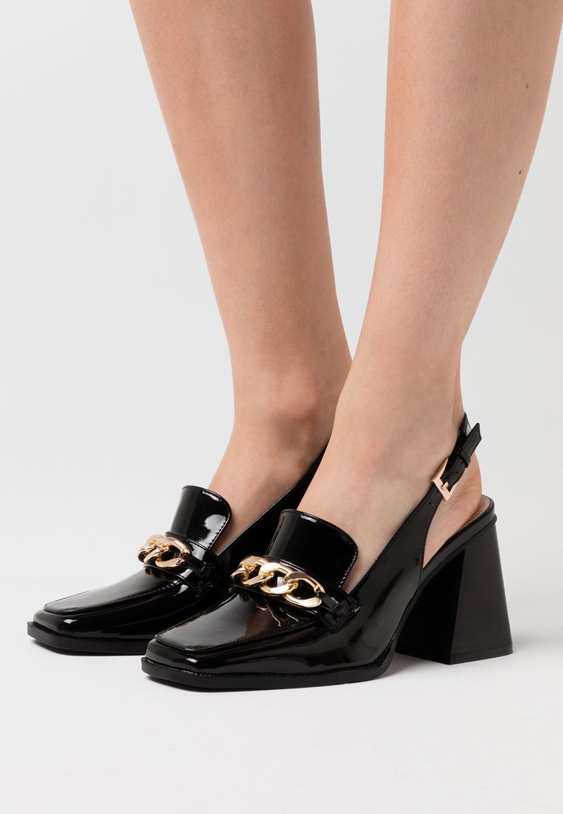 Escarpins noirs en cuir verni à bride arrière, talons blocs, bouts carrés et détail chaîne dorée sur le devant, portés pieds nus sur un fond neutre.