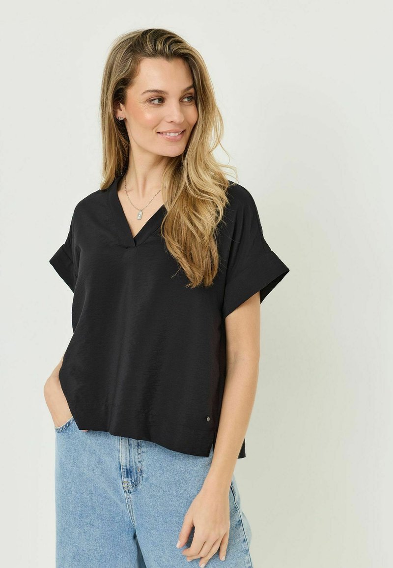 Femme aux longs cheveux blonds portant un haut noir à manches courtes et col en V ainsi qu'un jean bleu clair, souriante et regardant sur le côté.