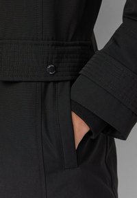 Zwarte jas met een textuurstof, een ceintuur in de taille en geribbelde manchetten. Opvallende details zijn de gestikte accenten en een logoknoop.