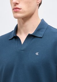 Νέος άνδρας που φοράει μπλε πόλο μπλουζάκι Calvin Klein με ανοικτό γιακά, εμφανιζόμενος από τον λαιμό έως το μέσο του στήθους σε απλό φόντο.