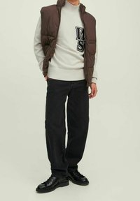 Person som bär en brun vadderad väst över en vit tröja med svarta bokstäver, svarta vida byxor och svarta snörskor.