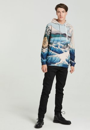 Hooded sweatshirt med et blåt og beige bølgemønster, med snor og lange ærmer. Båret med sorte bukser og støvler.