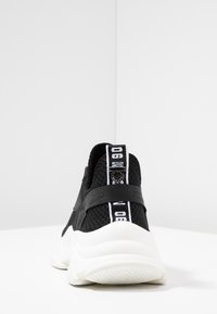 Zapatillas negras de punto con suela de goma, con un tirador blanco en el talón y una tira negra con un logo blanco en la parte trasera.