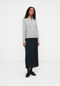 Suéter de punto gris con cremallera y mangas largas, combinado con una falda maxi plisada azul marino y zapatos negros, sobre un fondo blanco.
