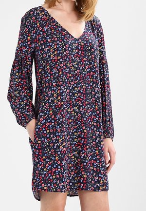 Vrouw draagt een marineblauwe bloemenjurk met lange mouwen en een V-hals, handen gedeeltelijk in de zijzakken, staand tegen een effen achtergrond.