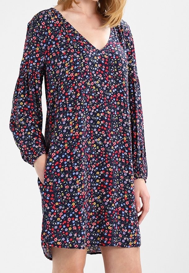 Femme portant une robe bleu marine à fleurs avec manches longues et col en V, les mains partiellement dans les poches latérales, debout devant un fond uni.