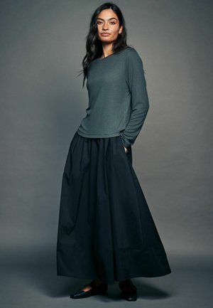 Långärmad, mörkgrön texturerad topp som matchas med en högmidjad, flödande svart kjol. Svarta platta skor kompletterar outfiten.