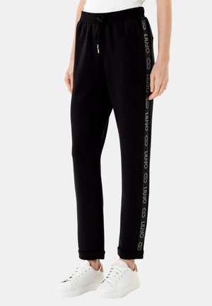 Pantaloni felpati neri con dettagli logo laterali, vita elastica, cordino e caviglie a costine, abbinati a sneakers bianche.