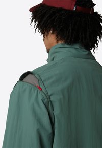 Grüne, leichte Jacke mit hohem Kragen und Reißverschlusstasche auf der Schulter, mit roten Akzenten und grauem Innenfutter.