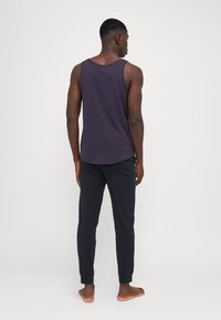 Débardeur violet à col ras du cou, associé à un pantalon noir fuselé avec des poignets élastiques. Tissu lisse ; porté pieds nus, vu de dos.
