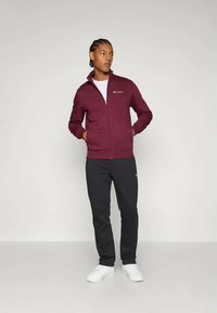 Bordeaux kleurige zip-up jas met logo, witte t-shirt eronder, gecombineerd met zwarte broek en witte schoenen. Glad textuur en een slim-fit ontwerp.