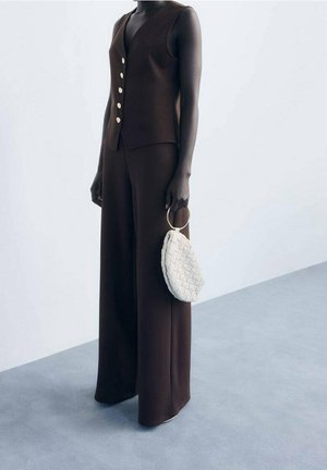 Modelo con un chaleco marrón sin mangas con botones dorados y pantalones de pierna ancha a juego, sosteniendo un bolso blanco tejido con un asa circular.