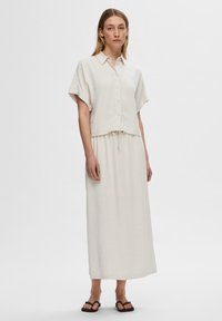 Light beige, short-sleeve, button-up shirt with a relaxed fit paired with a matching ankle-length skirt featuring an elastic waistband. Sandals complete the look.