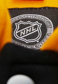 Ein schwarzes Stoffetikett zeigt das NHL-Logo, weiße Schrift auf einem strukturierten Hintergrund. Ein leuchtend gelber Teil des Bekleidungsstücks ist oben sichtbar.