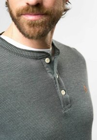 Pilkos vaflinės tekstūros ilgomis rankovėmis marškiniai su Henley apykakle, dviem kreminių spalvos mygtukais ir oranžiniu logotipu ant krūtinės. Grublėto audinio ir kontrastinga apykaklė.