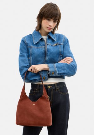 Noor naine sinises teksajakis ja mustades teksapükstes, hoides pruuni nahast käekotti ühe õlarihmaga.