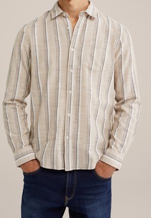 Homme portant une chemise beige rayée boutonnée avec une poche poitrine et un jean bleu foncé, mains dans les poches, debout devant un fond uni.