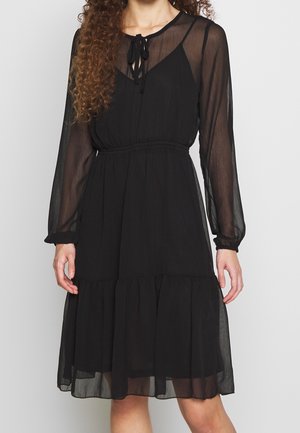 Donna che indossa un vestito lungo a maniche lunghe nero trasparente con scollatura a fiocco e vita arricciata, in piedi davanti a uno sfondo neutro.