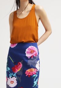 Top senza maniche marrone abbinato a una gonna blu aderente a fiori caratterizzata da grandi fiori rosa e bianchi, che mette in mostra colori vivaci e dettagliati motivi.