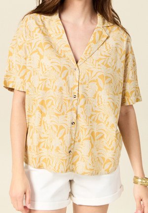 Vrouw draagt een geel overhemd met korte mouwen en witte botanische print, gecombineerd met witte shorts met opgerolde zoom, en een gouden armband als accessoire.