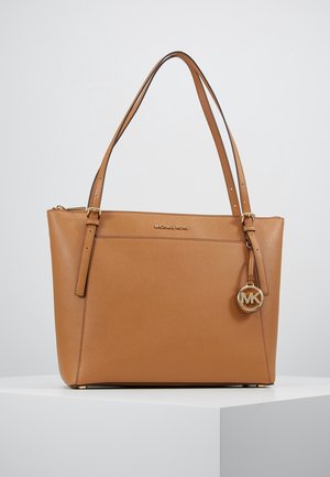 Tanfarvet læder Michael Kors shopper taske med dobbelte skulderremme, guldfarvet logo vedhæng og lynlåslukning på toppen, placeret på hvid piedestal.