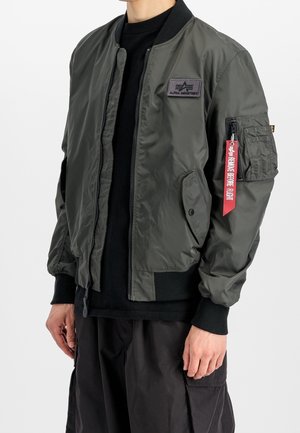 Personne portant une veste bomber Alpha Industries vert foncé avec des poignets et un col noirs, et une étiquette rouge "Remove Before Flight" sur la poche de la manche gauche.
