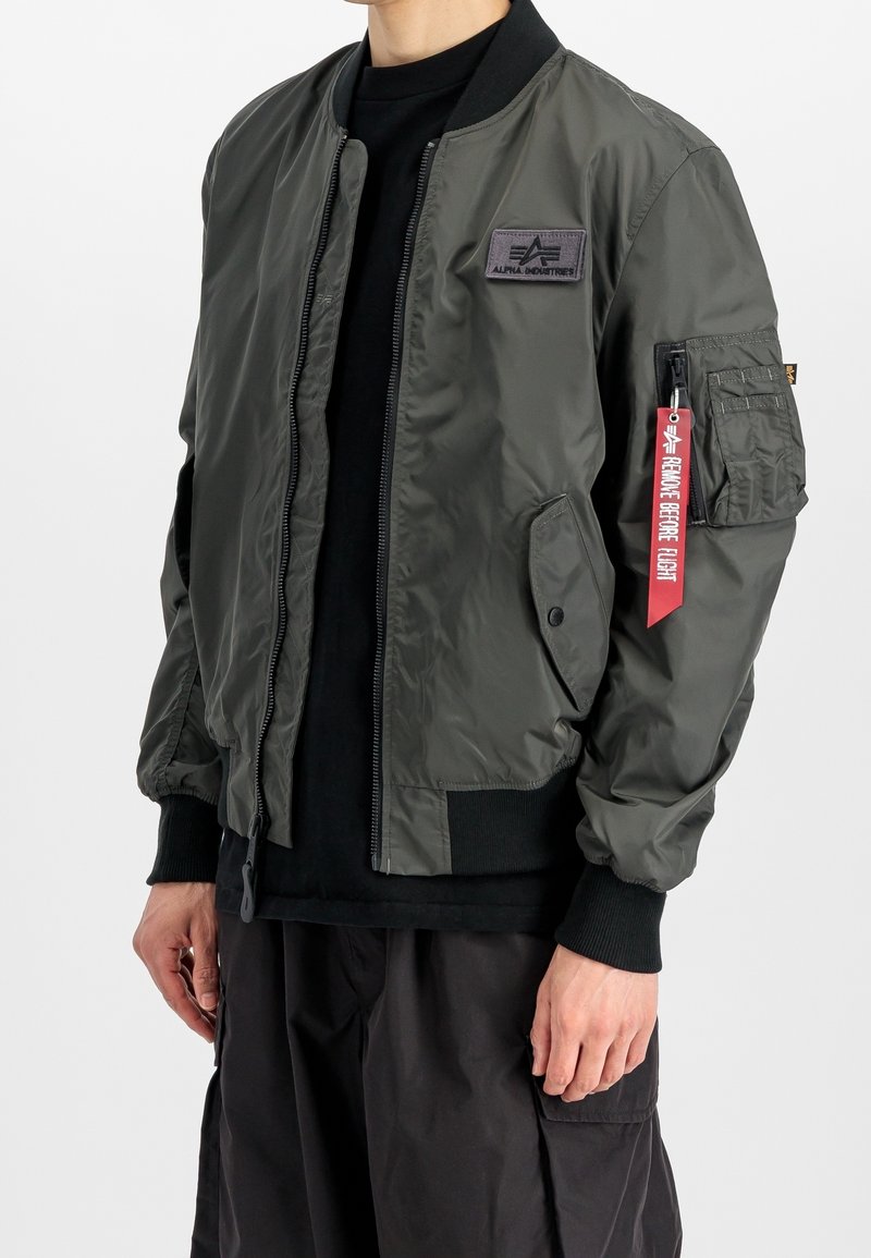 Personne portant une veste bomber Alpha Industries vert foncé avec des poignets et un col noirs, et une étiquette rouge "Remove Before Flight" sur la poche de la manche gauche.