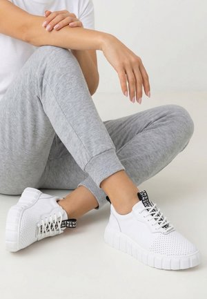 Person iført grå joggingbukser og hvide platformsneakers med sorte trækflapper, siddende med benene krydset på et lyst gulv.