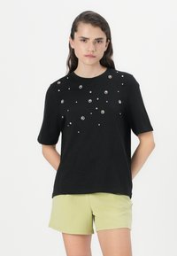 Zwarte T-shirt met korte mouwen en verspreide parelversieringen, ronde halslijn en een relaxte pasvorm, gecombineerd met lichtgroene shorts.