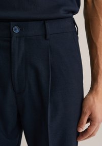 Marinblå byxor i texturerad tyg, med knäppning, framvik och sidofickor. Skräddarsydd passform som syns i handplaceringen.