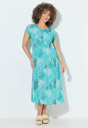 Kobieta z kręconymi włosami, ubrana w niebieską midi sukienkę w wzory i beżowe sandały na platformie, stojąca na białym tle.