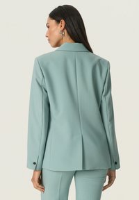 Világoskék, fazonra szabott zakó, sima textúrával és hosszú ujjakra tervezve, hátul szellőzővel, fekete gombdíszítéssel az ujjak végén.