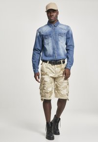 Denimskjorta med långa ärmar, ljusblå med två bröstfickor; beige camouflagemönstrade cargo shorts, svart bälte och svarta snörboots; beige keps.