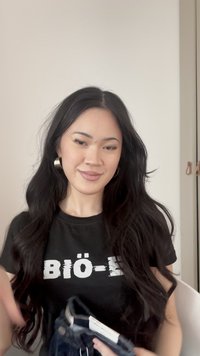 Jauna sieviete ar gariem tumšiem matiem valkā melnu krekliņu ar tekstu, turot salocītus džinsus rokās, stāvot pret vienveidīgu gaišu sienas fonu.