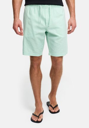 Mężczyzna w miętowo-zielonych spodenkach z troczkami i czarnych klapkach, stojący na białym tle.