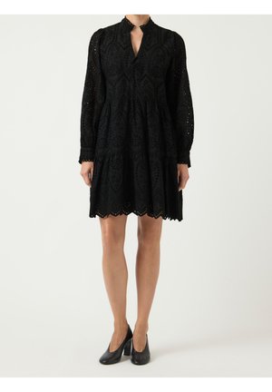 Kobieta w czarnej koronkowej sukience do kolan z długimi rękawami i wysokim kołnierzem, w połączeniu z czarnymi butami na obcasie, stoi na białym tle.