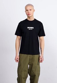 Camiseta de algodón negra de manga corta y cuello redondo, con el texto blanco "DICKIES" en la parte frontal. Combinada con pantalones cargo verdes.