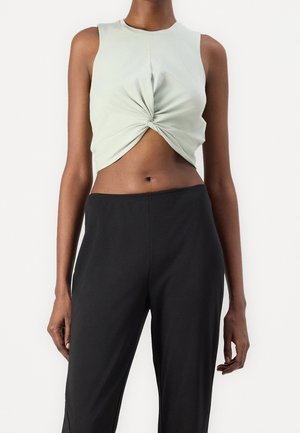 Osoba ubrana w bezrękawnik bladoniebieskiego koloru z skręconym węzłem crop top oraz czarne spodnie z wysokim stanem na jednolitym białym tle.