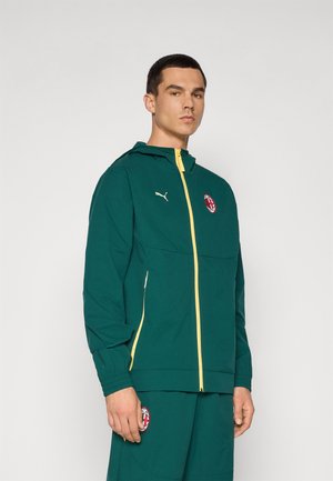 Teal jakke med gul lynlås, der har hætte og elastiske manchetter. Inkluderer et rødt og hvidt logo på venstre bryst og sidelommer.
