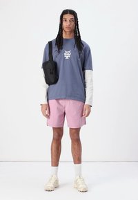 Uformell antrekk med en blå t-skjorte med grafisk design, rosa shorts og kremfargede joggesko, komplettert med en liten svart skulderveske.